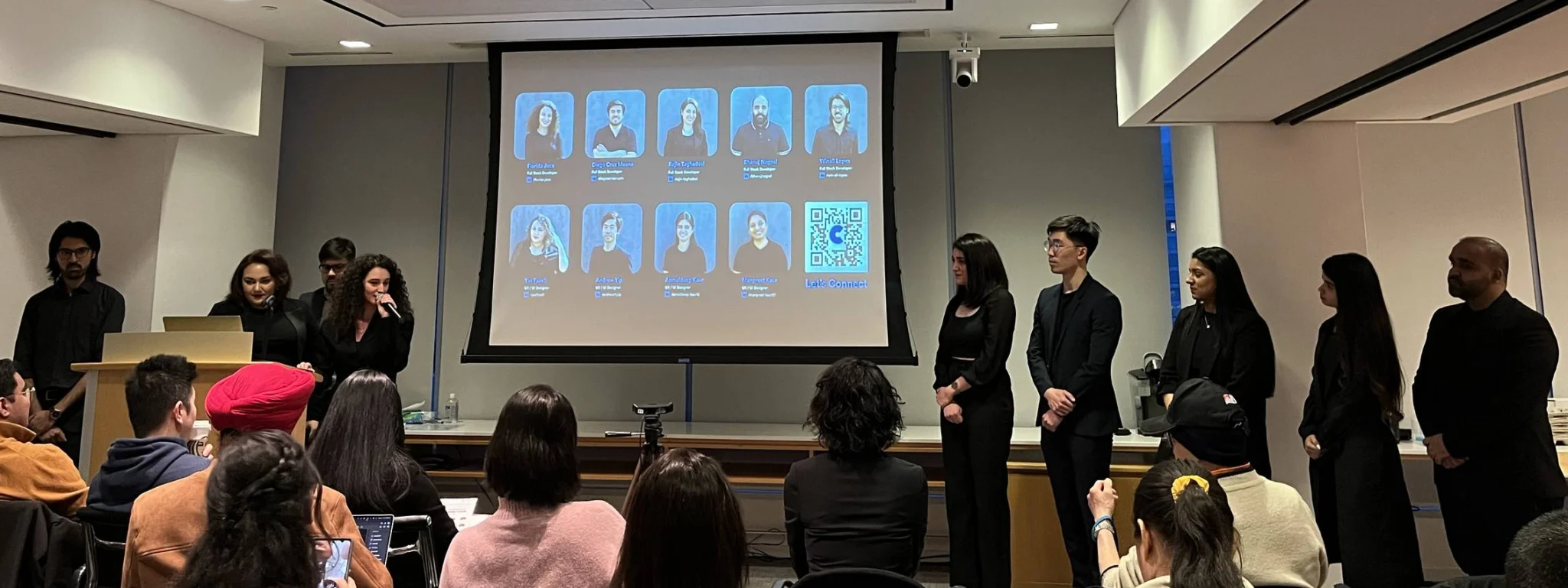 The Crowd Ease team presenting their project to a panel and audience, with a slide showing team member headshots and a QR code to connect
